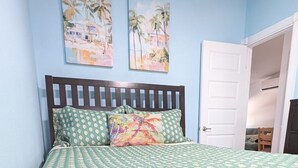 Une chambre à coucher avec un lit en bois, une couvre-lit à motifs verts, deux oreillers et deux tableaux représentant des palmiers, accrochés au mur.