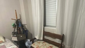 Ein kleines Zimmer mit einem Bett, einem Holzstuhl, einem Fenster mit Vorhängen, einem Ventilator und einem gelben Fitnessball.