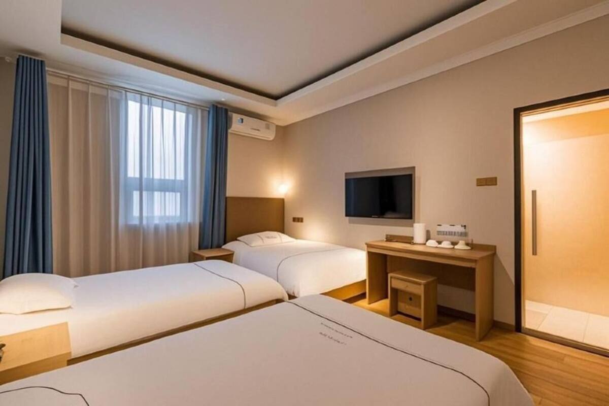A hotel room with two beds, a desk, a TV, and an air conditioning unit.