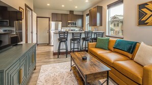 A modern kitchen with dark cabinets, a central island with stools, a wooden coffee table, and a leather sofa with cushions. - NEW! Town Center Basecamp at Big Sky | Walk to Dining, Shops & Events | Prime Location (Big Sky)