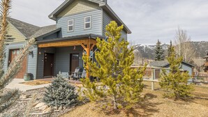 A two-story house with a porch, a chimney, and a small garden with pine trees. - NEW! Town Center Basecamp at Big Sky | Walk to Dining, Shops & Events | Prime Location (Big Sky)