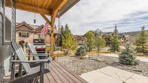 A wooden deck with Adirondack chairs, a grill, and a patio area with a view of neighboring houses and trees. - NEW! Town Center Basecamp at Big Sky | Walk to Dining, Shops & Events | Prime Location (Big Sky)