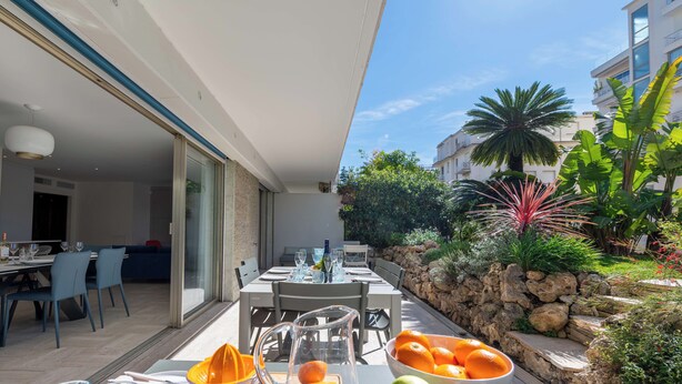 An outdoor dining area with a table set for two, a fruit bowl, and a stone wall with plants. - 3-Br Garden-Level Apartment – Near Beach with Parking (Cannes)