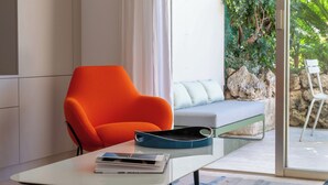 A modern room with a white table, a bright orange chair, a grey sofa, and a view of greenery outside. - 3-Br Garden-Level Apartment – Near Beach with Parking (Cannes)