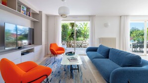 A modern living room with a flat-screen TV, orange and blue sofas, a coffee table, and a view of the outdoors. - 3-Br Garden-Level Apartment – Near Beach with Parking (Cannes)