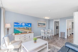 A modern living room with a flat-screen TV, a dining area with white chairs, a kitchen in the background, and a grey couch. - Ocean Dunes Tower I - Unit 4054 (Myrtle Beach)