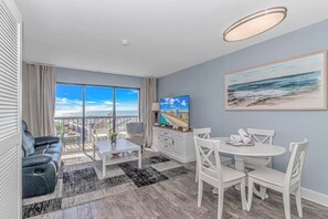 A modern living room with a balcony, a dining area, and a large window with a city view. - Ocean Dunes Tower I - Unit 4054 (Myrtle Beach)