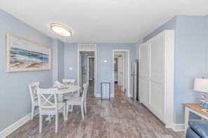 A room with a dining table, chairs, a painting, a trash bin, and a refrigerator. - Ocean Dunes Tower I - Unit 4054 (Myrtle Beach)