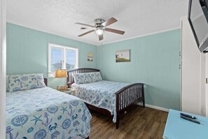 A bedroom with two beds, a ceiling fan, a window with blinds, and a small table with a lamp. - Blushing Mermaid (North Myrtle Beach)