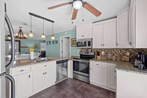 A kitchen with white cabinets, stainless steel appliances, a ceiling fan, pendant lights, and a backsplash. - Blushing Mermaid (North Myrtle Beach)