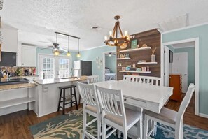 A kitchen with a white island, bar stools, a chandelier, and a dining table with chairs. - Blushing Mermaid (North Myrtle Beach)