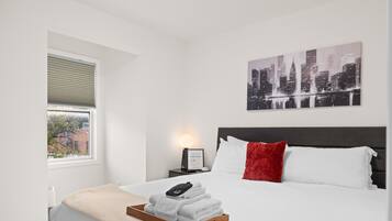 A neatly made bed with white linens and a red pillow, a wooden tray with folded towels, a framed picture of a cityscape, and a bedside lamp.