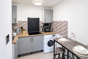 Et kompakt køkken med vaskemaskine, kaffestation og spiseplads med barstole.
