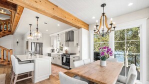 A spacious kitchen with white cabinetry, a wooden dining table, and a large window with a view of trees. - Bella Vista & Coastal Chateau: Lakefront Retreat with Dock, Kayaks, Bikes & Fire Pit (Seacrest)