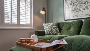Un canapé vert agrémenté d’un coussin à motifs de cactus, un plateau avec des tasses et une théière, et une œuvre d’art représentant un arbre, encadrée, accrochée au mur.
