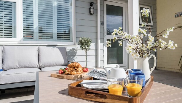 Un coin petit-déjeuner aménagé sur une terrasse en bois, avec un canapé, un plateau contenant des croissants, des fraises, une théière, des verres et un pichet de jus d’orange.