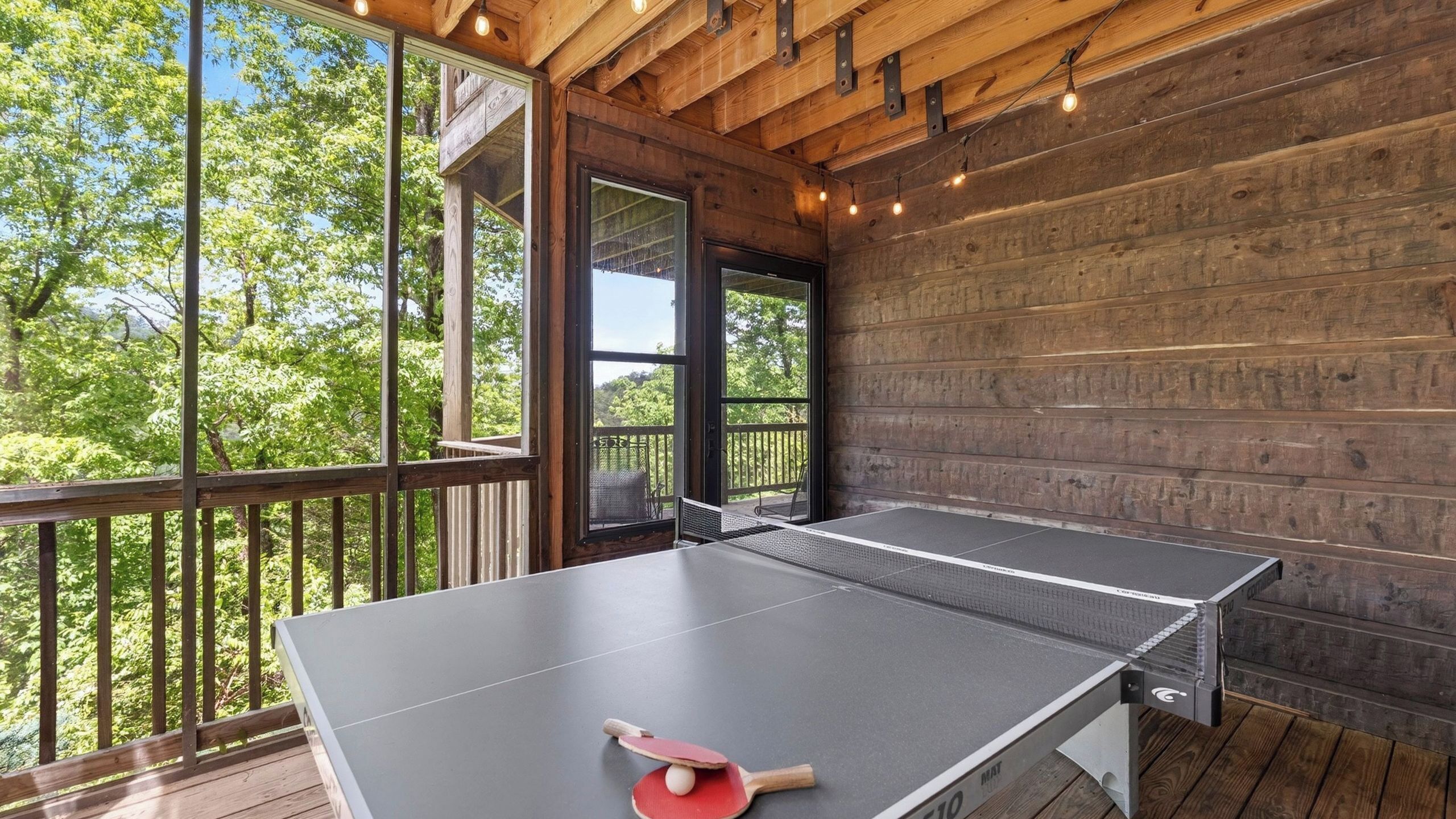 Une table de ping-pong avec une raquette et une balle, posée sur une terrasse en bois dotée de rampes en verre, offrant une vue sur une forêt.