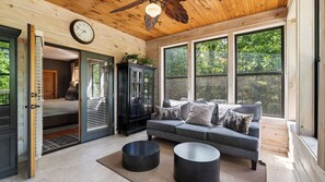 A cozy room with a grey sofa, two black round tables, a wooden ceiling fan, and large windows offering a view of trees. - Rocking Chair Ridge by Avantstay Lux Lodge Hot Tub + Fire Pit + Game Room + Yoga Room + Mountain Views (Sevierville)