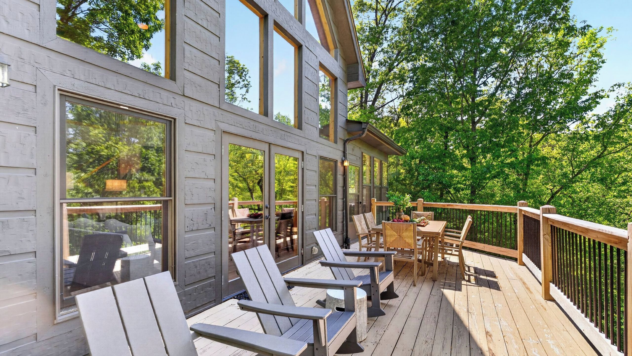 Una terraza de madera con mobiliario exterior, ventanas amplias y vista a árboles.