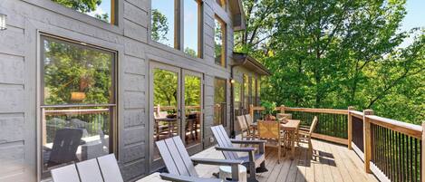 Una terraza de madera con mobiliario exterior, ventanas amplias y vista a árboles.