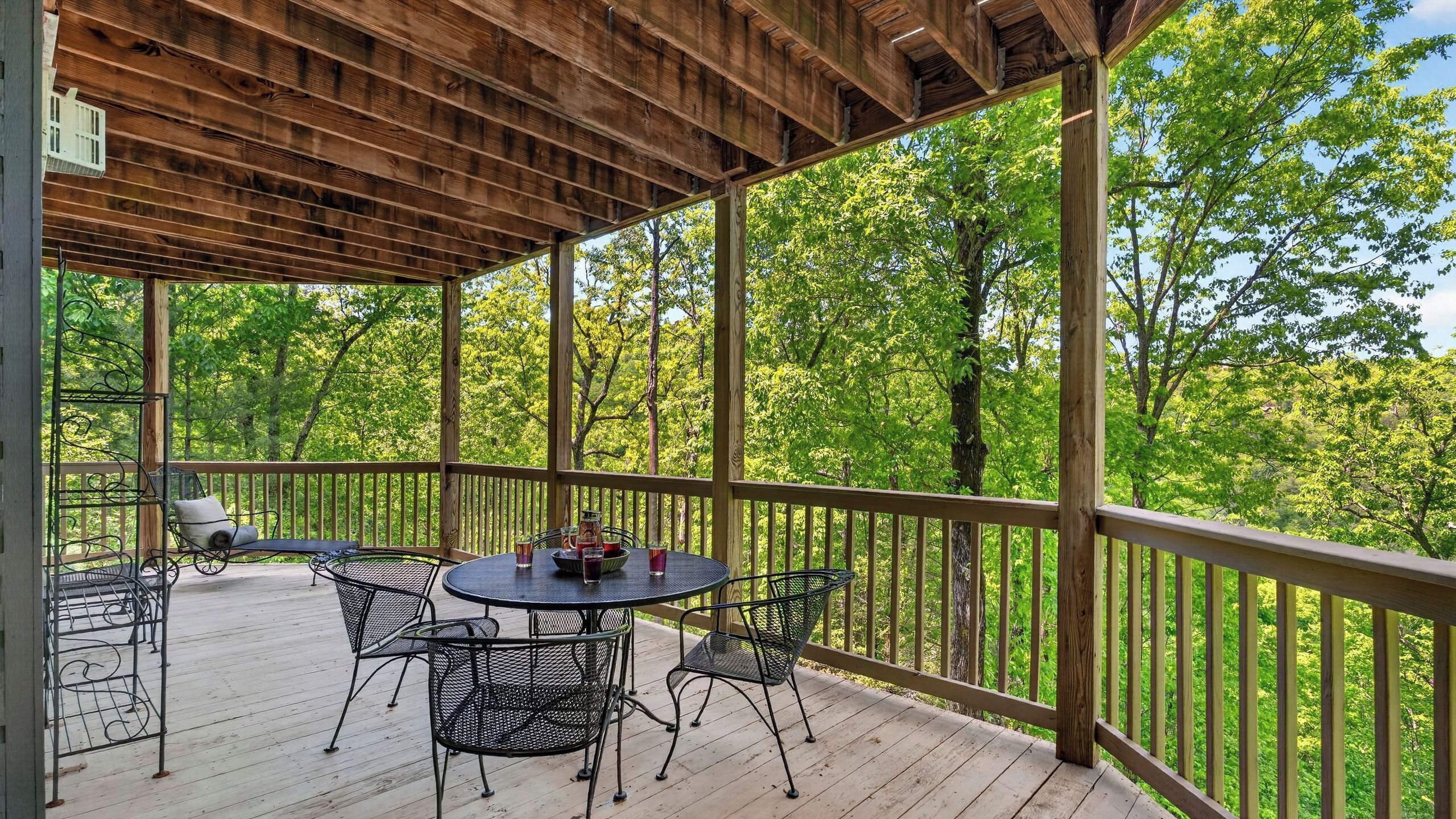 Une terrasse extérieure couverte, dotée d’une table et de chaises, entourée d’une rambarde en bois, et offrant une vue sur une zone boisée.