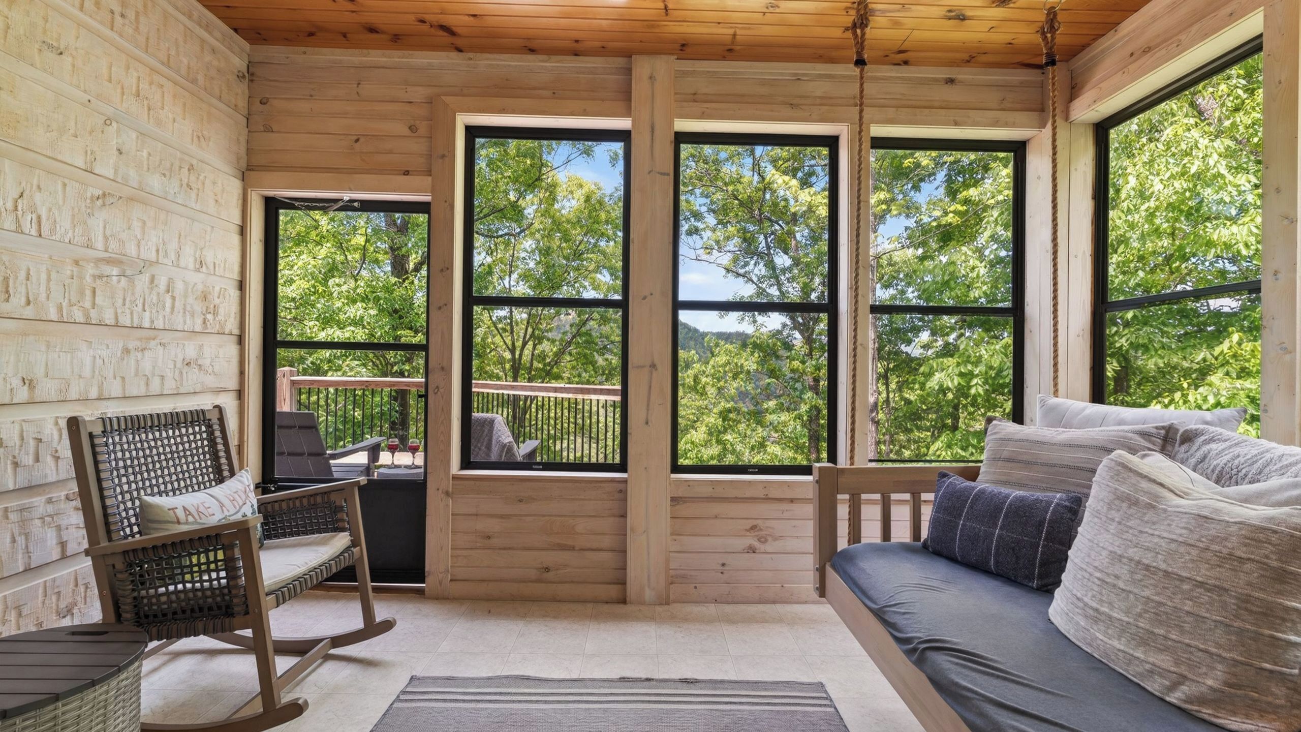 Een gezellig chalet interieur met een houten terras, een schommelstoel, een bank en grote ramen met uitzicht op bomen.