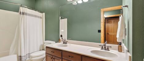 A bathroom with a double sink, a mirror, a shower curtain, and a toilet.