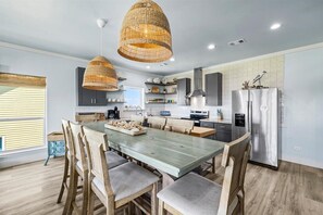 Eine moderne Küche mit Essbereich, ausgestattet mit einem großen Holztisch, Korbschwinglampen und einem Edelstahlkühlschrank.