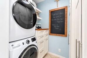 A laundry room with a front-loading washer and dryer, a chalkboard with a Bible verse, and a wooden shelf with decorative items. - Modern 3BR Galveston with Fire Pit & Private Pool & Jacuzzi | Mins to the Beach (Galveston)