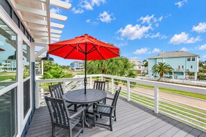 A balcony with a round table and four chairs, a red umbrella, and a view of houses and greenery. - Modern 3BR Galveston with Fire Pit & Private Pool & Jacuzzi | Mins to the Beach (Galveston)