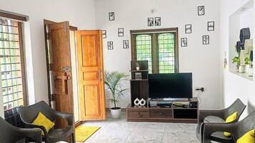 A spacious living room with a flat-screen TV, two sofas with cushions, a ceiling fan, a wooden door, and a large window with a view.