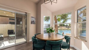 Un coin repas avec une table ronde, six chaises, une plante en pot et une vue sur une piscine et des palmiers.