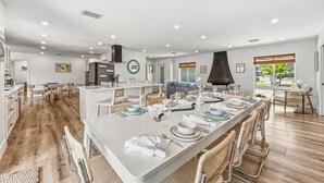 Un'ampia cucina con elettrodomestici moderni, un'area pranzo con un lungo tavolo apparecchiato per sei persone e un soggiorno con divano e una grande finestra.