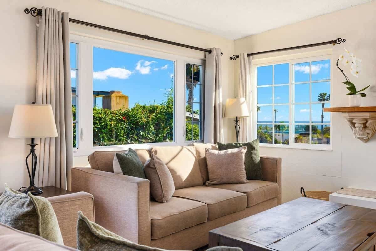 Quarto com sofá bege, duas luminárias, mesa de centro de madeira e grandes janelas com cortinas.