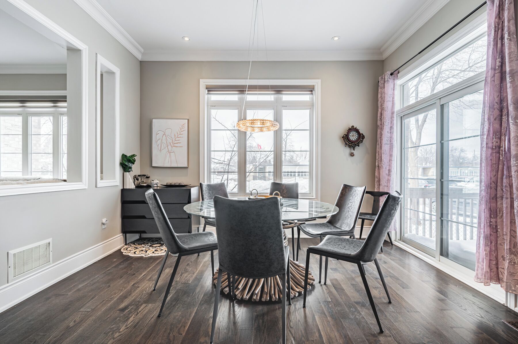 A dining room with a round table, six chairs, a chandelier, and large windows with curtains.