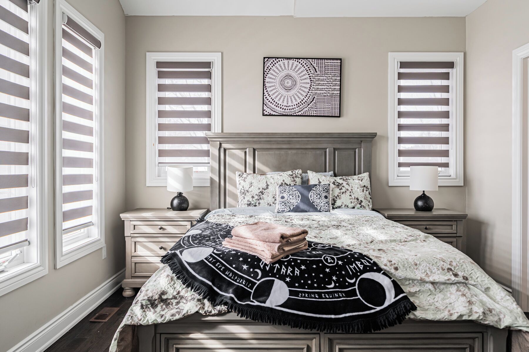 A bedroom with a bed, two bedside tables, a headboard, and two windows with blinds.