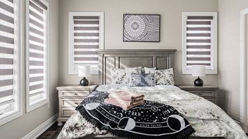 A bedroom with a bed, two bedside tables, a headboard, and two windows with blinds.