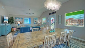 Un coin repas avec une table en bois et des chaises en osier, un ventilateur de plafond et une vue sur le quartier grâce à de grandes fenêtres.