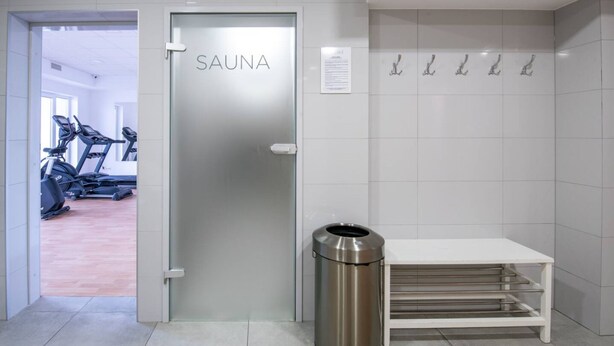 Un interior moderno con una puerta que lleva a la sauna, señalizada con 'SAUNA', un cubo de basura y un banco blanco.