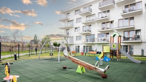 Un parque infantil con sube y bajas y columpios, rodeado por un edificio blanco con balcones.