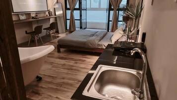A modern hotel room with a large bed, a bathtub, a sink, and a view of the street through large windows.