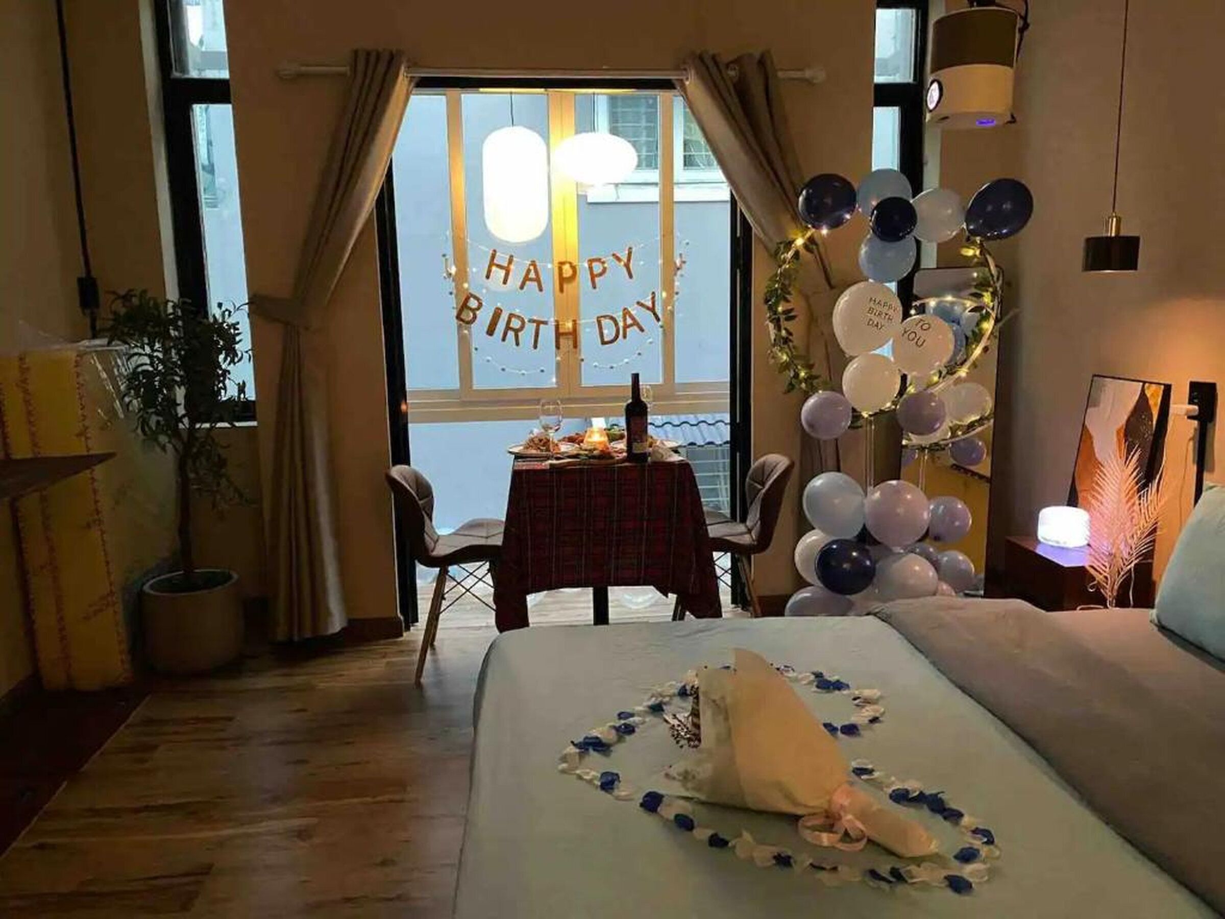 Habitación de hotel con cama, una mesa con botella y vasos, una ventana con un cartel de 'HAPPY BIRTHDAY' y globos.