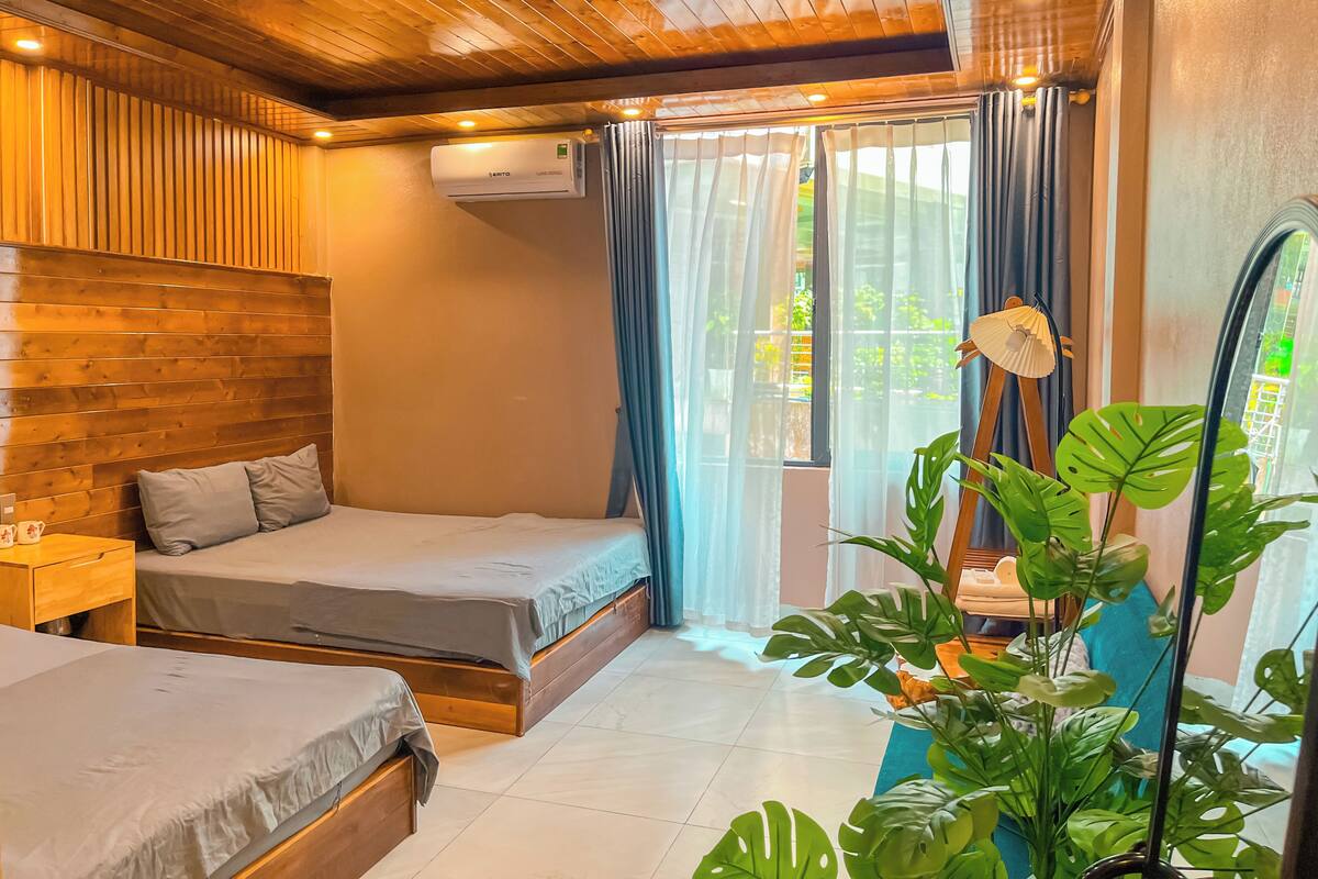 Ein Zimmer mit Holzboden, einem Bett mit grauer Matratze, einem Nachttisch, einem Fenster mit Vorhängen und einer Zimmerpflanze.