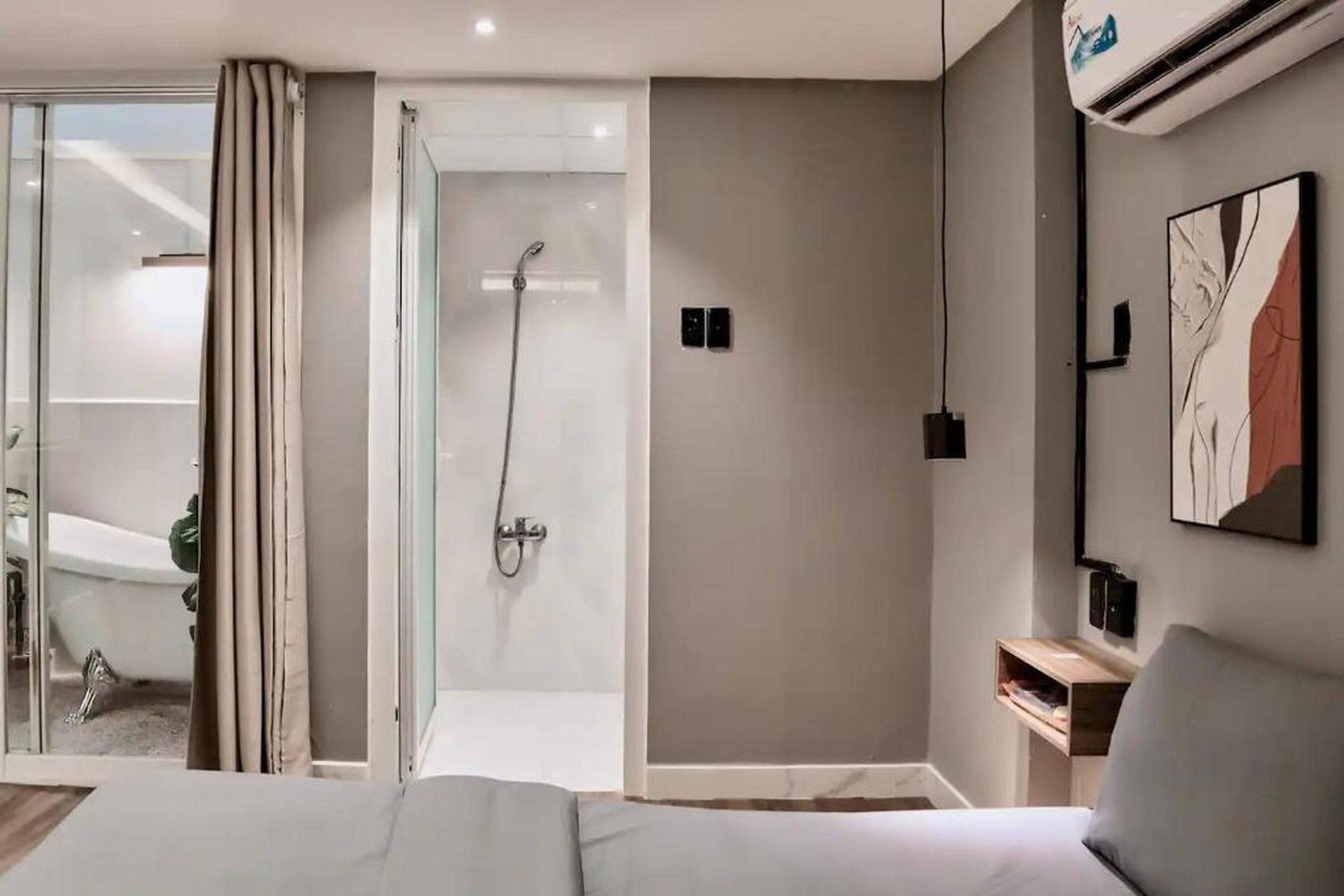 A modern hotel room with a glass shower enclosure, a bed, a bedside table, and a wall-mounted air conditioning unit.