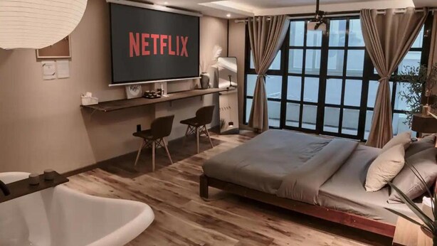 Un dormitorio moderno con una cama grande, una pantalla de televisor mostrando 'NETFLIX', una bañera y un ventanal con vistas.