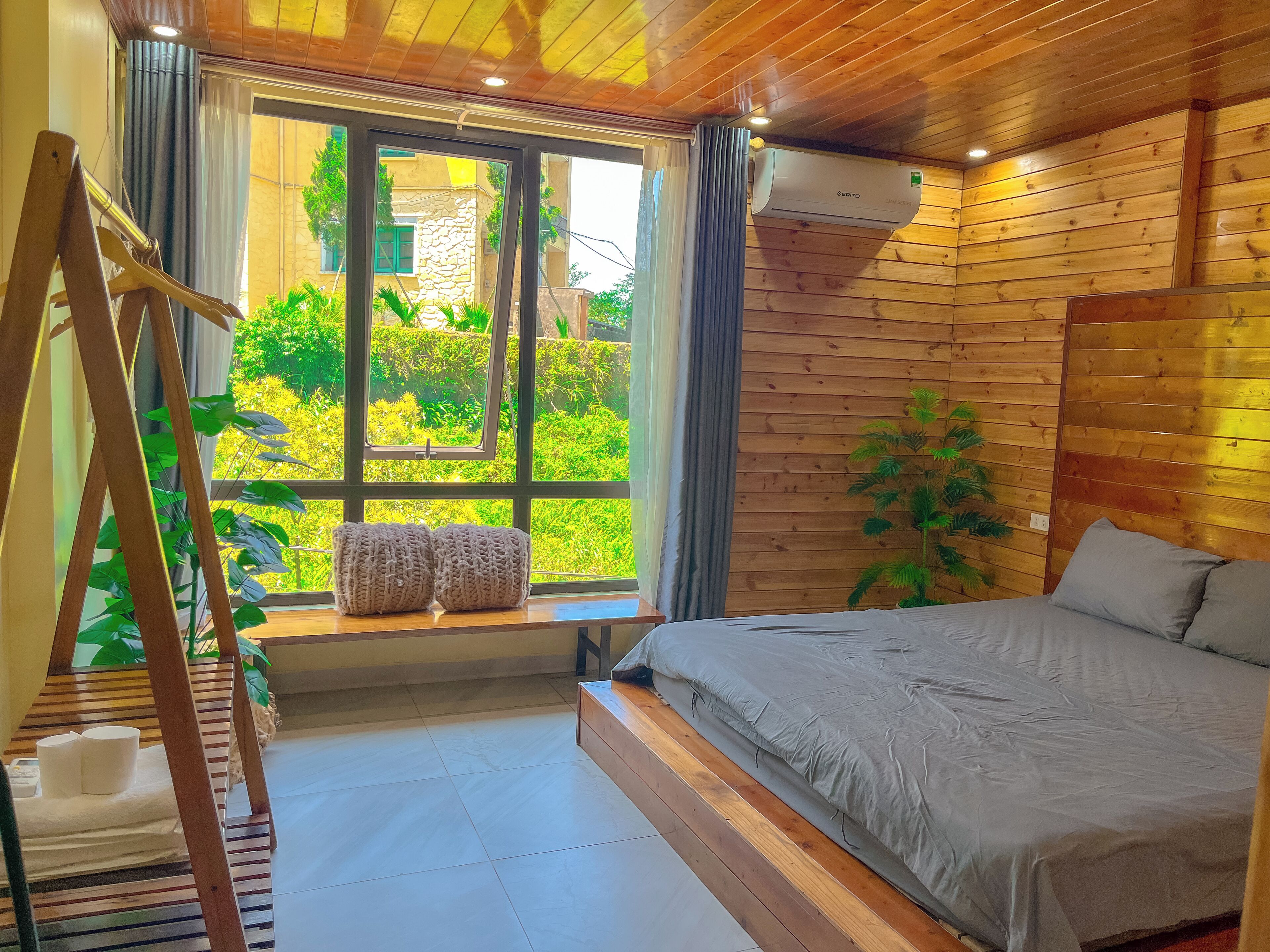 A bedroom with a bed, a window with curtains, a wooden ceiling, and a view of greenery outside.