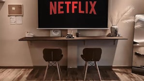 Habitación con una televisión de pantalla grande mostrando 'NETFLIX', un escritorio de madera con dos sillas, un jarrón con plantas secas y un cuadro en la pared.