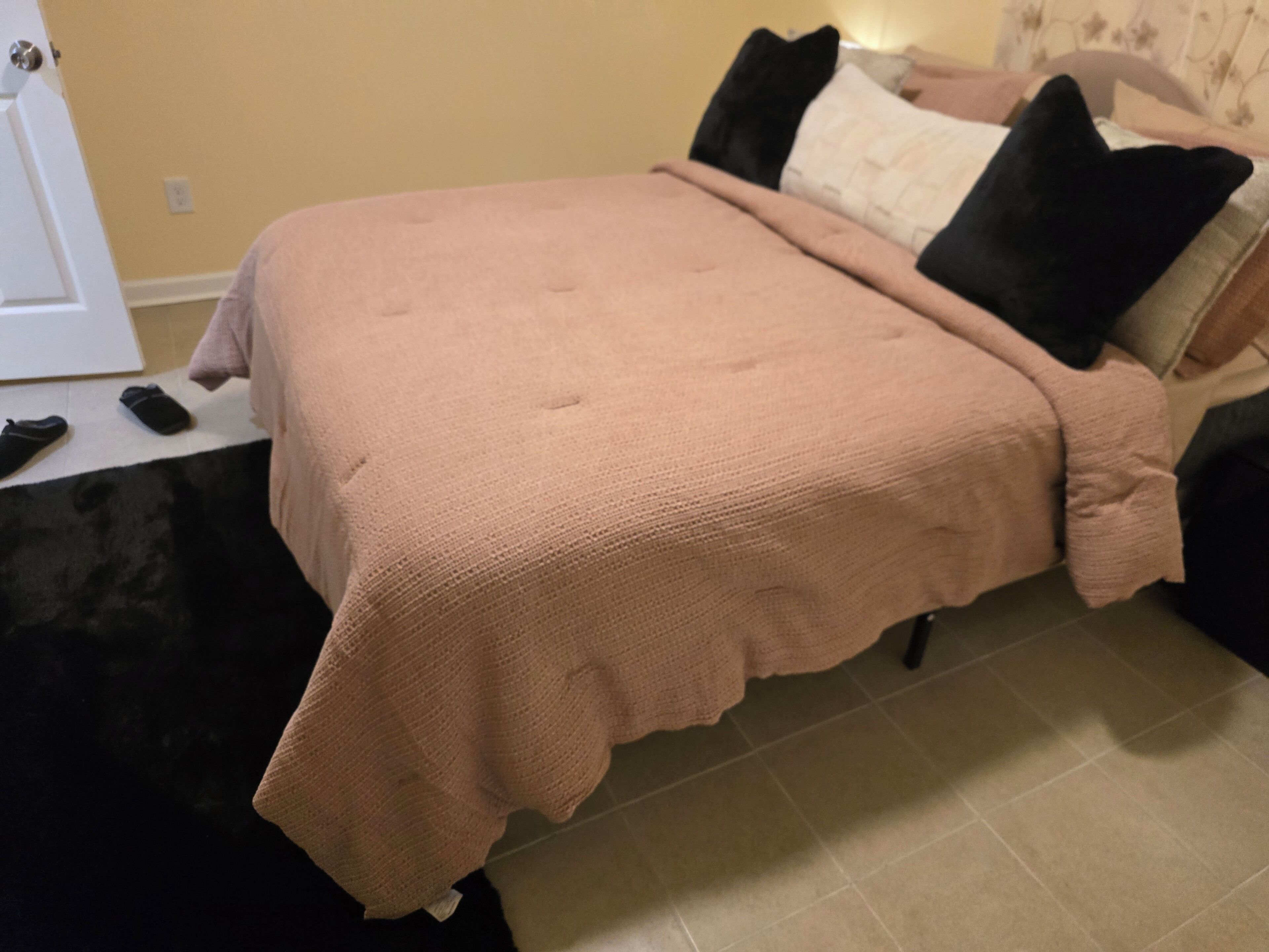 A bed with a pink blanket, black and white pillows, and a remote control on a black surface.