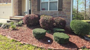 A residential house with a brick facade, a front porch, and a well-maintained garden with mulch and shrubs.