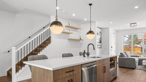 Cozinha moderna com ilha branca, máquina de lavar louça em aço inoxidável e iluminação pendente.
